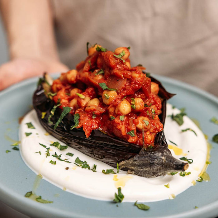Two hands holding a plate with a baked eggplant covered in spicy braised chickpeas on Greek yogurt sprinkled with chopped mint.