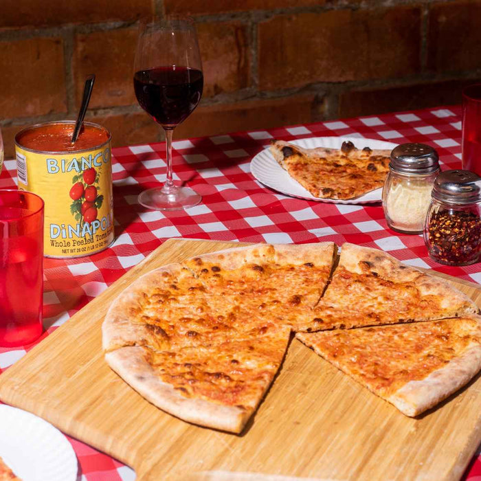 New York-style Käsepizza auf einem Ooni Pizzaschieber und Pizzabrett aus Bambus auf einem Tisch neben Pizzastücken, einer Dose Bianco di Napoli-Tomaten und Gläsern. 