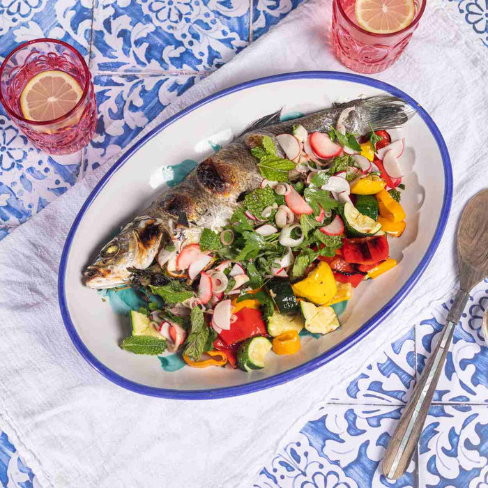 Whole roasted branzino with squash, peppers, shaved spring onions, fennel, radish, parsley, dill and tarragon on a plate on a table.