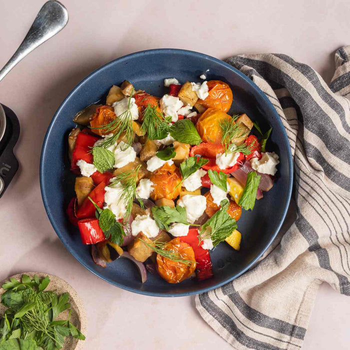 Langsam gerösteter Kürbis, Aubergine, Zwiebeln, Kirschtomaten und Paprika in einer Ooni Ofenform neben einer Schüssel mit langsam geröstetem Gemüse mit Feta und frischen Kräutern auf einem Tisch. 